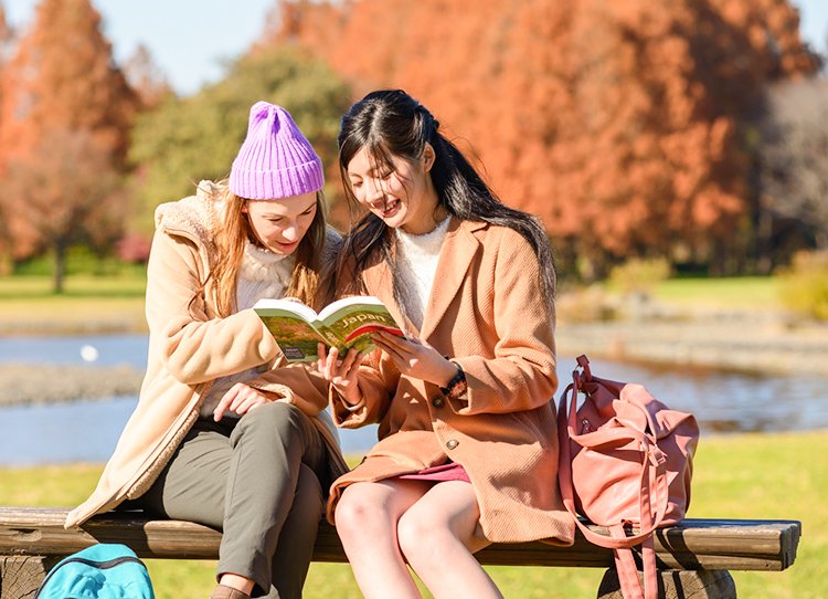 公園のベンチで日本のガイドブックを見る日本人女性と外国人女性